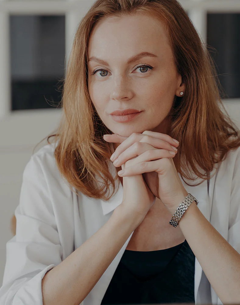 A light-skinned woman with shoulder-length reddish-brown hair is seated, looking directly at the camera with a thoughtful or serious expression. Her light-colored eyes are focused, and her hands are clasped in front of her chin. She wears a white long-sleeved button-up shirt over a dark top, with a silver-toned watch on her right wrist. The softly lit background is out of focus, featuring off-white walls or a light-colored room. - Eyelid Surgery in Newport News, VA
