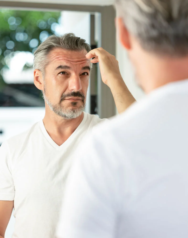 A middle-aged man with gray hair and a trimmed beard looks at his reflection in a mirror. Wearing a plain white v-neck t-shirt, he touches his forehead with one hand, appearing concerned or thoughtful. The blurred background suggests a bathroom. His reflection shows the back of his head and shirt. - Fue Hair Transplant in Newport News, VA